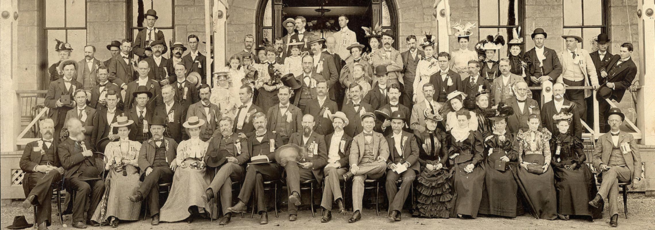 Photo of a group of men and women from 1898 wearing period clothes and arranged in a group outside a building.