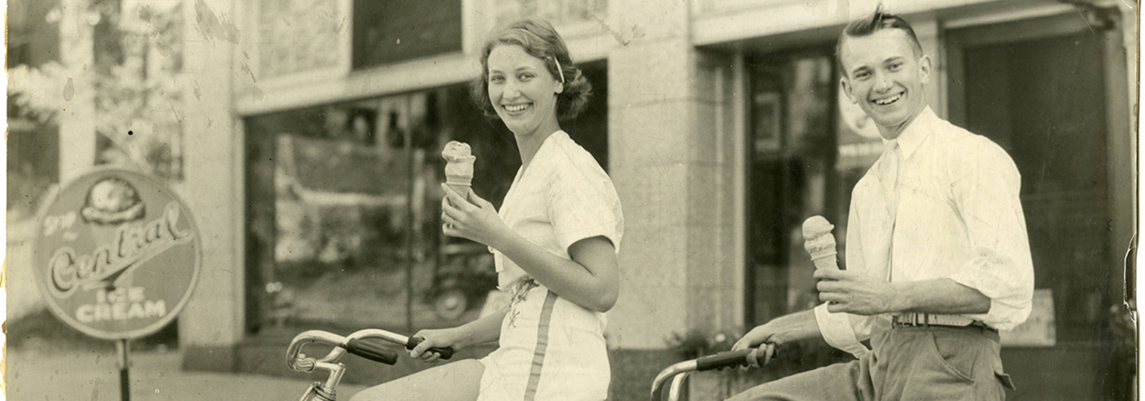 A woman and a man on a tandem bicycle holding Central Dairy ice cream cones and smiling at camera